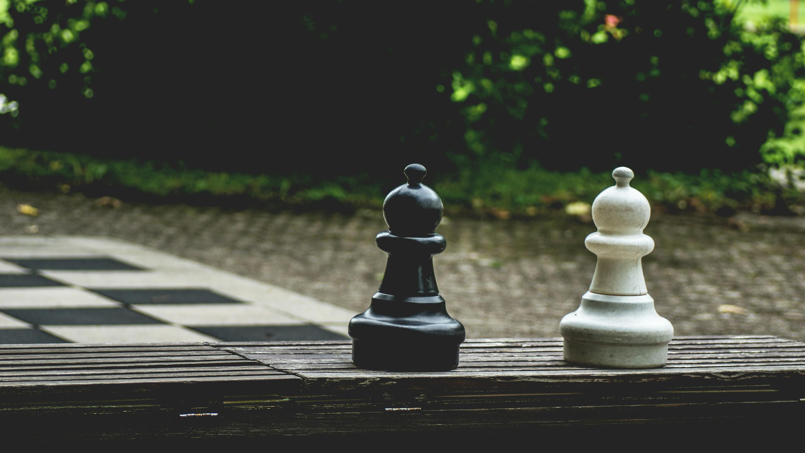 Close-up of large black and white chess pieces on a wooden bench outdoors, symbolizing strategy.